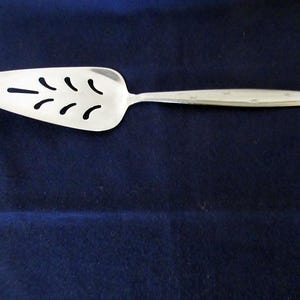 Puede incluir: Un servidor de pastel plateado con una hoja triangular ranurada y un mango largo y delgado. La hoja tiene una serie de recortes curvos. El servidor está hecho de un metal brillante y reflectante sobre un fondo azul oscuro.