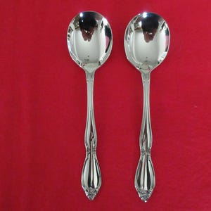 May include: Two silver soup spoons with oval bowls and ornate handles are displayed against a red background. The spoons are highly polished, reflecting the surrounding environment. The handles have a decorative floral design at the base.