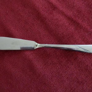 Pode incluir: Uma faca de manteiga de aço inoxidável com uma lâmina larga e arredondada e um cabo curvo. A faca está colocada sobre um fundo vermelho escuro, destacando sua superfície polida e seu design elegante. Este utensílio é perfeito para espalhar manteiga ou queijos macios.