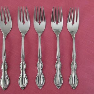 Può includere: Cinque forchette d'argento con manici decorati sono disposte su uno sfondo rosa. Ogni forchetta ha quattro rebbi e un manico decorativo con un design dettagliato. Le forchette sono ben distanziate e riflettono la luce.