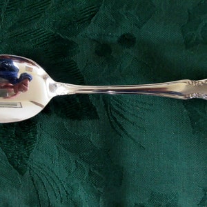 Può includere: Un cucchiaio d'argento con una ciotola arrotondata e un manico decorato. Il cucchiaio è su una tovaglia verde.