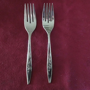 May include: Two stainless steel forks with a floral design on the handles. The forks have four tines and are set against a burgundy background. The forks are approximately 20 cm long.