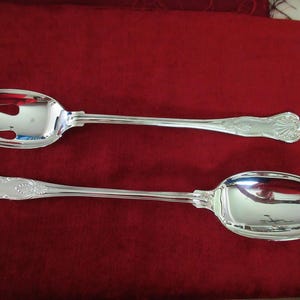 Könnte beinhalten: Ein silbernes Serviergabel- und Löffel-Set. Die Gabel hat drei Zinken, und der Löffel hat eine große, ovale Schüssel. Beide Utensilien haben verzierte Griffe mit einem Muscheldesign. Das Set wird auf einer roten Oberfläche präsentiert.