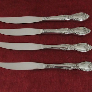 May include: Four silver dinner knives with ornate handles. The knives have a polished finish and are arranged in a row on a red surface.
