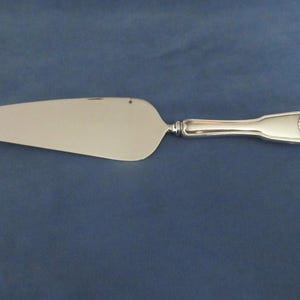 May include: A silver-toned cake server with a wide, flat, triangular blade and a smooth, polished finish. The handle features a rounded end with a decorative embossed shell motif at the top, providing a classic look for serving desserts.