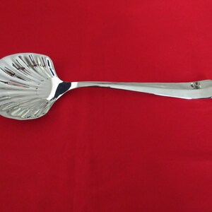 Puede incluir: Una cuchara de servir de plata con un cuenco en forma de concha. La cuchara tiene un mango largo y delgado.