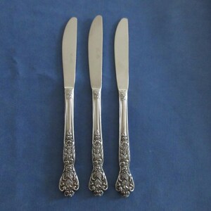 Può includere: Tre coltelli da burro argentati con decorazioni floreali sui manici sono disposti verticalmente su uno sfondo blu. I coltelli hanno lame curve e motivi dettagliati, suggerendo uno stile vintage o antico. I manici presentano intricati disegni floreali.