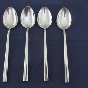 May include: A set of four identical silver-toned metal spoons arranged in a vertical row. Each spoon features a smooth, oval-shaped bowl and a long, straight, flat handle with a polished, reflective finish that catches the light.