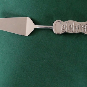 May include: A silver-colored cake server with the word "Celebrate" on the handle. The server has a wide, flat blade and a long, thin handle. The background is a solid, dark green color.