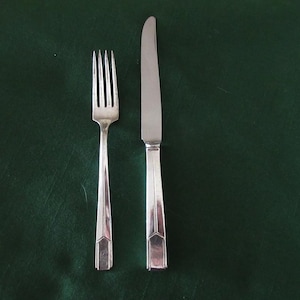 May include: Silverware set featuring a fork and knife with geometric handle details. The utensils are placed on a dark green surface, creating a classic and elegant aesthetic. The fork has four tines, and the knife has a smooth, sharp blade.