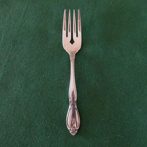 Può includere: Una forchetta d'argento con quattro rebbi e un manico decorativo. La forchetta ha un ritaglio a forma di diamante tra i rebbi. Il manico ha un motivo floreale alla base. La forchetta è posta su uno sfondo verde scuro.