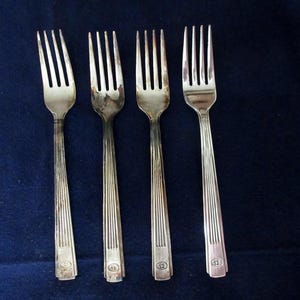 Może przedstawiać: Cztery srebrne widelce z długimi, żłobkowanymi trzonkami i czterema zębami każdy, ułożone na ciemnoniebieskiej powierzchni. Widelce mają prosty, elegancki design, odpowiedni do formalnych posiłków. Każdy widelec ma mały emblemat u podstawy trzonka.