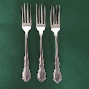 May include: Three silver forks with ornate handles are arranged on a dark green surface. Each fork has four tines and a decorative, elongated handle. The forks are made of a shiny, reflective metal.