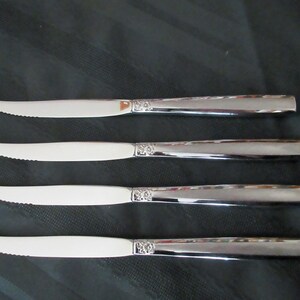 Könnte beinhalten: Vier silberfarbene Steakmesser mit gezahnten Klingen. Die Messer haben ein dekoratives Blumendesign am Klingensockel und einen glänzenden, reflektierenden Griff. Die Messer sind horizontal auf einer dunklen Oberfläche angeordnet.