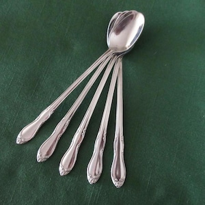 Può includere: Un set di cinque cucchiai d'argento con manici decorati, disposti su una superficie verde scuro. I cucchiai presentano un design classico con un motivo floreale dettagliato sui manici e una finitura lucida e riflettente.