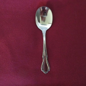 Puede incluir: Una cuchara plateada brillante con un cuenco ovalado y un mango decorativo. La cuchara se encuentra sobre un fondo rojo oscuro, resaltando su superficie pulida y su intrincado diseño. Una pieza clásica de cubertería.
