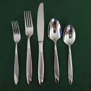 Könnte beinhalten: Ein Set aus fünf Edelstahl-Besteckteilen, darunter zwei Gabeln, ein Messer und zwei Löffel. Das Besteck hat ein schlichtes, elegantes Design mit einem diagonalen Ätzdetail an den Griffen. Die Teile sind auf einer dunkelgrünen Oberfläche angeordnet.