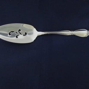 May include: A silver-colored pie server with a slotted, oval-shaped head and a decorative handle. The server has a floral design at the end of the handle. The background is a dark blue.