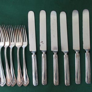 May include: A set of six silver-toned metal forks and six silver-toned metal knives arranged in two rows. Each piece features a slender handle with a decorative embossed pattern at the base and a polished finish on the tines and blades.