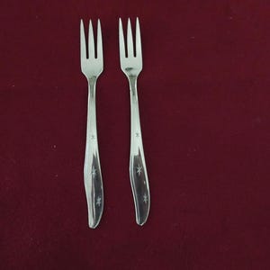 May include: Two silver forks, each with three tines, displayed on a deep red backdrop. The handles feature a curved shape and are adorned with small star-shaped engravings. The forks are arranged vertically, angled slightly towards each other.