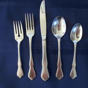 May include: A collection of five silver-coloured cutlery items displayed on a dark blue surface. The set includes two forks, a knife, and two spoons. The utensils have decorative handles and are arranged in a row.