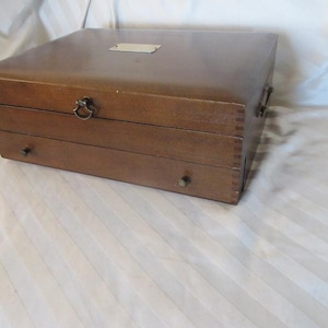 Puede incluir: Una caja de almacenamiento rectangular de madera marrón con tapa abatible, una pequeña placa de metal plateado, un pestillo central y un cajón inferior con dos pequeños pomos redondos. La caja presenta uniones de dedos visibles.