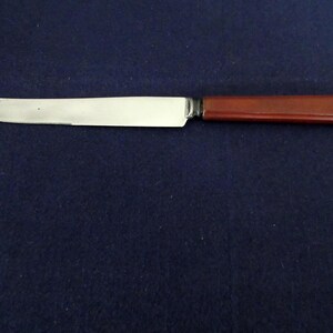 Könnte beinhalten: Ein Buttermesser mit einer glänzenden, silbernen Klinge und einem dunkelbraunen Griff. Das Messer ist vor einem dunkelblauen Hintergrund platziert, was seine schlichte Eleganz hervorhebt. Der Griff scheint aus Holz oder einem ähnlichen Material zu bestehen.