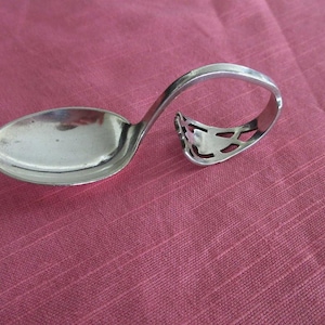 Puede incluir: Una cuchara de plata con un diseño de mango único. El mango se curva en un bucle con un recorte decorativo. La cuchara descansa sobre una superficie de tela rosa, mostrando su superficie pulida y reflectante.