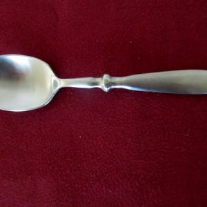 May include: A silver-toned metal spoon with a wide, rounded bowl and a smooth, tapered handle. The handle features a distinct, raised circular collar detail where it connects to the neck of the spoon, providing a simple and functional design.