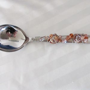 May include: A decorative spoon with a polished silver bowl and handle. The handle is adorned with wire-wrapped beads in shades of peach, pink, and clear. The spoon is designed for serving or display.