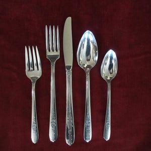 Op de afbeelding: Een set van vijf zilverkleurige bestekstukken, gerangschikt op een donkerrode achtergrond. De set bevat twee vorken, een mes en twee lepels. Elk stuk heeft een bloemmotief op het handvat.