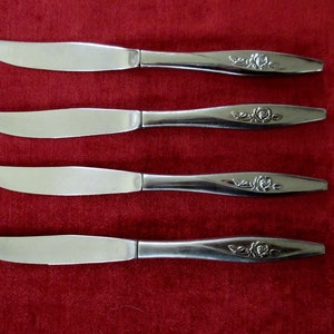 May include: Four silver dinner knives with rose designs on the handles. The knives are arranged in a row on a red surface.