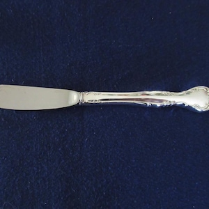 Può includere: Un coltello da burro argentato con una lama arrotondata e un manico ornato. Il manico presenta un design floreale dettagliato. Il coltello è posto su uno sfondo blu scuro, evidenziando la sua superficie lucida. Questo elegante utensile è adatto per cene formali.
