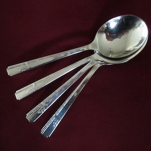 May include: Four silver soup spoons arranged on a dark red surface. The spoons have a classic design with a decorative pattern on the handles. The spoons are made of a shiny, reflective metal.