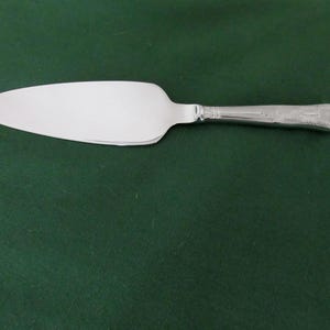 Könnte beinhalten: Ein silberner Tortenheber mit einer breiten, flachen Klinge und einem dekorativen Griff. Der Griff weist ein verziertes Design auf. Der Heber befindet sich vor einem dunkelgrünen Hintergrund.