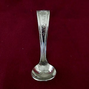 May include: A small, silver spoon with the word "cream" engraved on the handle. The spoon has a rounded bowl and a slender handle. The spoon is likely vintage and is set against a dark red background.