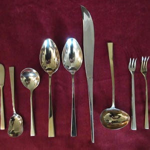 May include: A collection of polished silverware, including spoons, forks, knives, and a ladle, arranged on a deep red surface. The silverware has a shiny, reflective surface, with some pieces featuring gold-colored accents. The arrangement suggests a formal dining setting.