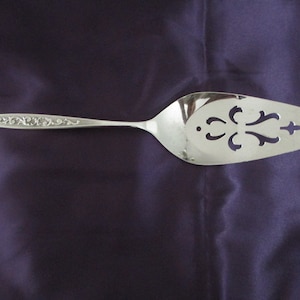 Pode incluir: Um servidor de bolo de prata com um design ornamentado e recortado. O cabo é decorado com um padrão floral.