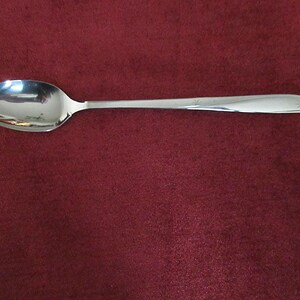 May include: A shiny, silver spoon with a rounded bowl and a long, slender handle. The spoon is set against a deep red background, creating a contrast that highlights its polished surface. The handle has a subtle decorative element near the end.