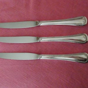 May include: Three stainless steel dinner knives with a decorative beaded design on the handles. The blades are smooth and the handles have a rounded end. The knives are arranged on a pink surface.