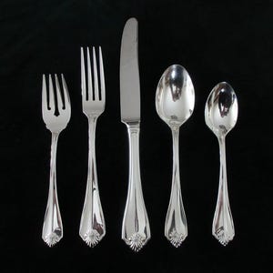 May include: A set of five silver-colored utensils arranged on a black surface. The set includes two forks, a knife, and two spoons. Each piece has a decorative floral design at the base of the handle. The utensils are reflective.
