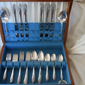 Könnte beinhalten: Ein Vintage-Besteckset in einer Holzkiste. Das Set enthält Löffel, Gabeln und Messer, alle mit verzierten Griffen. Der Innenraum des Koffers ist mit blauem Stoff ausgekleidet, was einen Kontrast zu den silbernen Utensilien bildet.