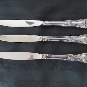 Peut inclure: Trois couteaux à beurre argentés avec des détails floraux ornés sur les manches. Les lames sont incurvées et reflètent l'environnement. Les couteaux sont disposés horizontalement sur un fond sombre, mettant en valeur leur design élégant.