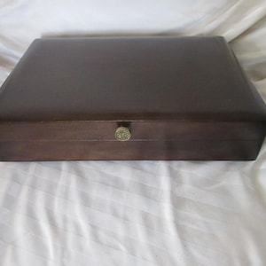 Puede incluir: Una caja rectangular de madera marrón oscuro con un pomo color latón. La caja está cerrada y se encuentra sobre una superficie blanca texturizada. La caja tiene un diseño simple y elegante.