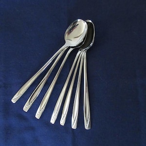 Puede incluir: Un juego de seis cucharas plateadas con mangos alargados y cuencos ovalados. Están dispuestas sobre una superficie azul oscuro, reflejando la luz. Los mangos presentan un sutil patrón decorativo cerca de la base.