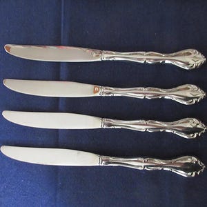 Può includere: Quattro coltelli da burro argentati con manici ornati sono disposti su una superficie blu scuro. Le lame sono lisce e i manici presentano un design floreale dettagliato. I coltelli sono un set, probabilmente per una cena formale.