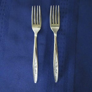 May include: Two silver-toned metal forks with four tines each. The handles feature a tapered design with a textured, recessed panel near the base that displays a subtle, embossed floral pattern. The forks have a polished finish on the upper stems.