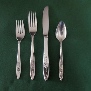 May include: A set of four silver-colored utensils: a fork, a knife, a spoon, and another fork. Each piece has a floral design on the handle. The utensils are arranged on a dark green surface.
