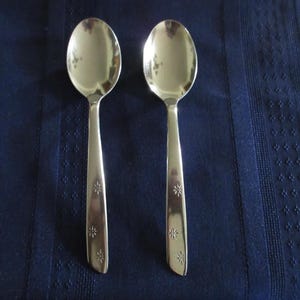 May include: Two silver spoons with oval bowls and long handles. The handles are decorated with a floral pattern. The spoons are set against a dark blue background.