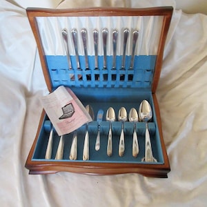 May include: A wooden cutlery chest with a hinged lid, featuring a blue velvet interior lining with slots for silver-toned spoons and forks. A small informational pamphlet with the text Prestige is placed inside the open case.